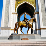 A majestic, high-resolution architectural photograph of the Ismail Somoni Monument in Dushanbe. The towering bronze equestrian statue of Ismail Somoni stands atop a grand, ornate stone pedestal adorned with intricate bas-reliefs depicting historical and cultural scenes. A prominent golden arch elegantly crowns the monument. The setting is Rudaki Park, with lush, well-maintained gardens and blooming flowers under a clear, serene morning sky, bathed in soft, natural light. Professional photography, epic scale, high detail, vibrant colors, safe for work, appropriate content, family-friendly.